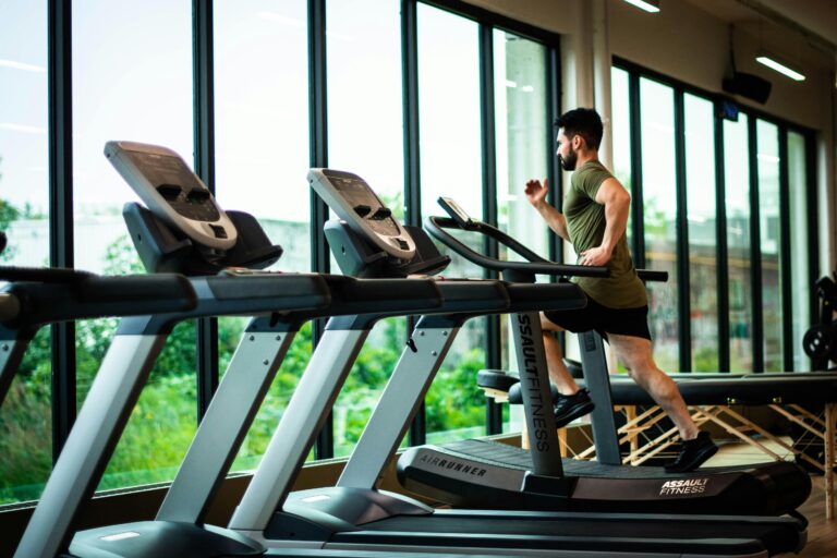 Man running on a treadmill inside a gym with large windows in the background